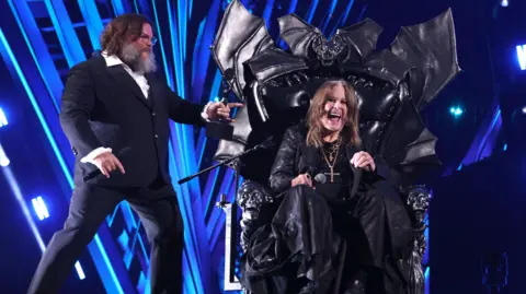 Getty Images The image shows two men. Jack Black is standing up. He is wearing a dark suit with a white shirt. He has shoulder-length brown hair and a beard. The second, Ozzy Osbourne, is still in large black leather bat-shaped throne. He is smiling. He had shoulder-length brown hair long dark clothes and a crucifix around his neck. They are in front of a blue and black backdrop. 