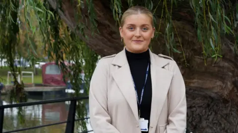BBC Elle Johnson is looking at the camera with a faint smile, she has a pale jacket on and black turtleneck jumper, and is wearing a lanyard. She is standing in front of a weeping willow beside the canal in Ely.