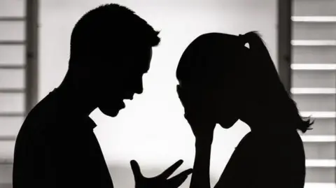 Getty Images A silhouette of a man and a woman facing each other, standing in front of a window. The woman has her head in her hands and looks upset and distressed, while the man is waving an open palm and seems frustrated and angry