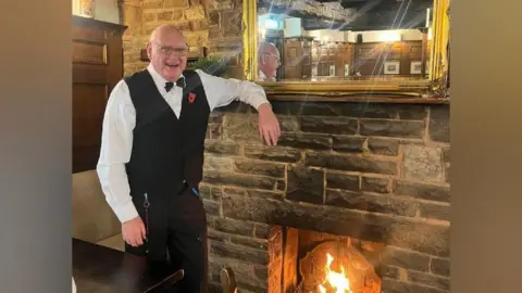 Amy Woodhouse Dennis Duckhouse, a bald man, stands leaning against a brick fireplace. He is wearing a black waistcoat and whit shirt, along with a black bow tie. On the wall behind him is a gold mirror. He is smiling