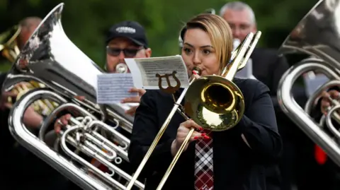 Reuters A woman playing a brass instrument. She is a member of a marching brass band which is playing in a parade during the traditional Whit Friday band contest in Manchester in 2024. Behind her can be seen two men who are also playing brass instruments