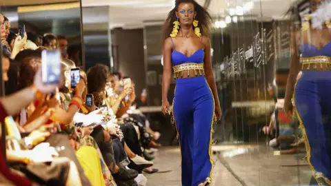 EPA A model walking down a catwalk during African Fashion International Fashion Week in Cape Town, South Africa as people in the crowd take pictures. The model is wearing clothes by UNICEF ambassador and South African designer Gavin Rajah.