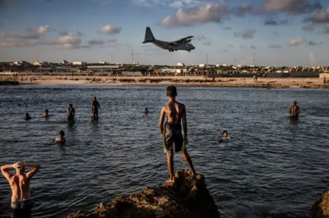 AFP Soldiers and residents swim next to Mogadishu's air base as a plane flies overhead.