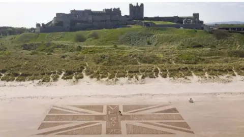PA Media A giant union flag created with footprints on the beach beneath Bamburgh castle in Northumberland