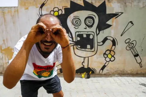 AFP Tunisian graffiti artist Kaisser Grojja, also known as "Wolf Gang", poses for a picture next to his latest mural in the town of Sidi Bouzid in central Tunisia on August 27, 2021
