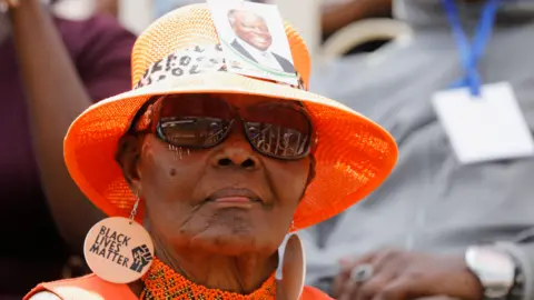 Reuters A woman in hat with Mwai Kibaki's image stuck to it and in Black Lives Matter earrings at Nyayo Stadium, Nairobi, Kenya - Friday 29 April 2022