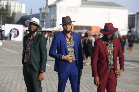 EPA Three men wearing suits, multicoloured shirts and hats are posing for a photo.