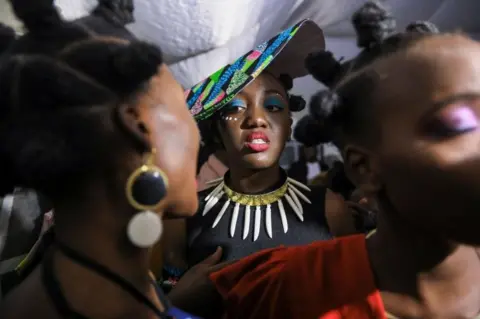 EPA A woman with elaborate face make up and a hat is pictured. In front of her there are two women with bantu knots.