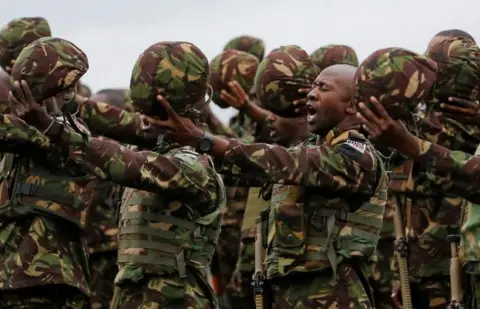 Reuters The Kenyan military wearing army uniform are holding their helmets in the air and appear to be singing.