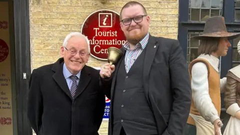 City of Ely Council Chris Phillips, City of Ely Mayor, is standing next to Matthew Routledge, the new town crier. Chris is wearing a black coat with a blue shirt and a striped tie. Matthew is wearing a black suit and a striped shirt. He is holding a gold bell. They are standing outside the Tourist Information Centre and Cromwell Museum