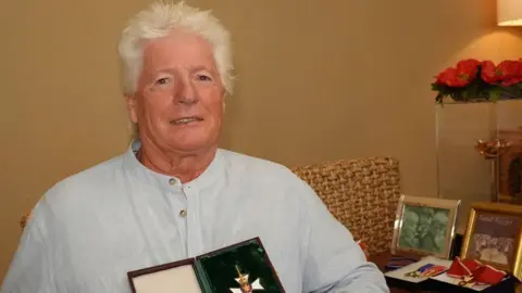 PA Media Bruno Peek who has white hair and is wearing a pale blue striped collarless shirt. He is sitting down and is holding up a case in which an order can be seen. There is a desk to his left with photos and more honours