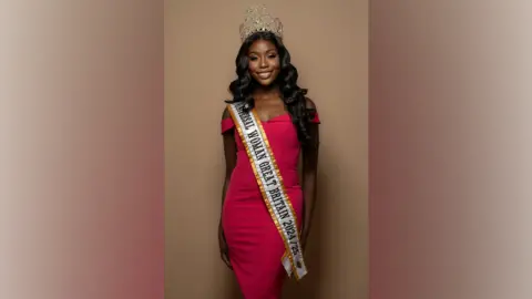 Martin Higgs A black woman with long black hair past her shoulders stands facing the camera. She wears a pink-red dress with a long sash which says "Universal Woman Great Britain 2024/25" on it. She has a tiara on her head.