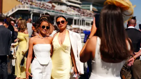 PA Media Two women in sunglasses pose arm-in-arm as another woman in a large hat takes their photo. The packed grandstand at Aintree forms the backdrop.
