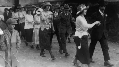 Helston Flora Day Association A black and white photograph of nine couples dancing in 1924. The men are wearing trilby hats and the ladies dark skirts with white blouses and all of them are wearing lily of the valley buttonholes. There is a young boy in the foreground wearing a flat cap and long coat.