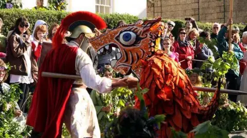 Helston Flora Day Association A man wearing a golden helmet with a red ruff and cape is jousting with a character dressed as a dragon wearing a papier mache head that has big teeth in its open mouth. They are outside and a crowd of spectators is gathered around them. 