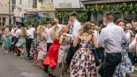 Discover Helston Young men and women are dancing in the street. The women's mid-length dresses are brightly coloured and many have floral designs. The women are wearing gloves. The men are wearing white shirts and black trousers with green and yellow striped ties. The shops lining the street are decorated with sunflowers, greenery and bluebells. There are spectators on the pavement.
