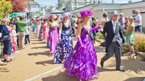 Helston Flora Day Association Couples are walking along a gravel walkway away from a street with bunting. The ladies are wearing brightly-coloured full-length gowns with hats and long gloves. The gentlemen are wearing top hats and tails. It is bright sunny day and the path is lined with spectators.