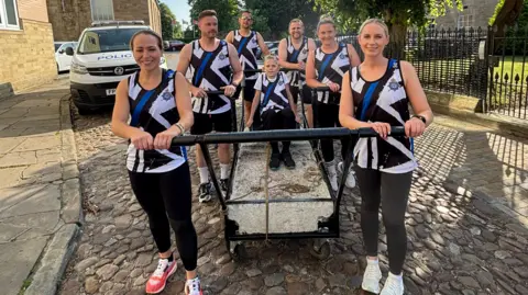 BBC /Sarah Urwin Six people (three women and three men) wearing white vests and black jogging bottoms, stand around a trolley on wheels. A small child wearin the same outfit sits on the trolley.