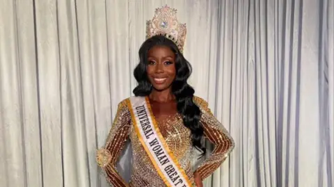 Natalie Mageza A woman wearing a large gold crown and a golden dress with a yellow and white sash which says "Universal Woman Great Britain". She is standing and smiling, with her hand on her hip, in front of a white curtain.