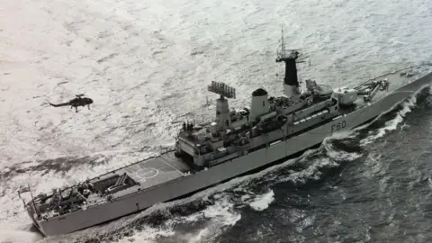 HMS Jupiter Association An aerial shot of HMS Jupiter. The black and white image shows a long, slim battleship at sea, with a helicopter flying over it.