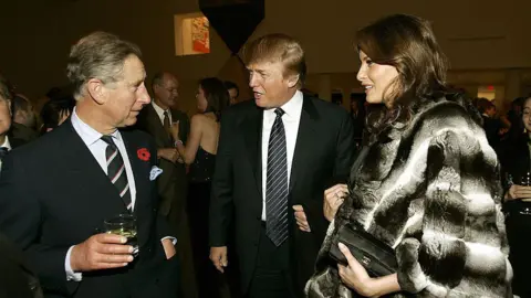 Getty Images A younger King Charles, wearing a red poppy lapel pin and holding a drink, chats to Donald Trump and Melania. Drump wears a dark suit, and Melania is wearing a big black-and-white fur coat and carrying a black Chanel clutch