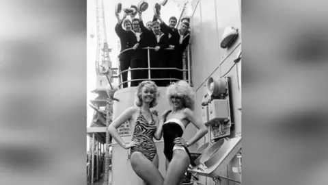 HMS Jupiter Association Two beauty contest entrants on the deck of the HMS Jupiter during a visit in 1991. The two women are wearing swimming costumes and are smiling for the camera, holding one hand on their hip and the other hand on each other's shoulder. Behind them is a group of men in uniform, waving hats.