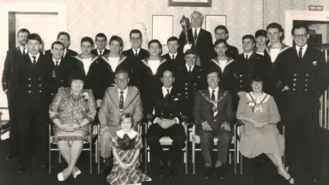 HMS Jupiter Association A black-and-white photograph taken at Middlesbrough town hall, showing members of the crew led by the Captain Cdr Colin Hamilton with civic dignitaries including Middlesbrough Mayor Malcolm Kirk and Mace Bearer Bernard Martin. The crew are wearing uniforms, while the mayoral party is wearing formalwear and chains.
