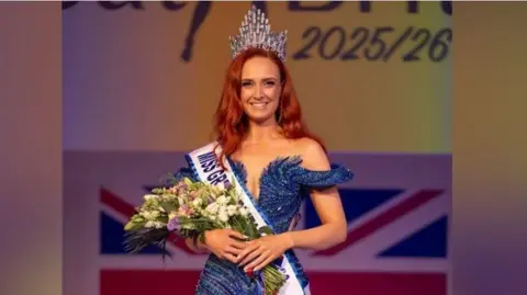 Chris Perfect Alice Cutler wearing a dark blue sparkly dress, a sash with the words: "Miss Great Britain" and holding a bunch of flowers and a crown in her hair. She has red hair and is stood on stage after winning.