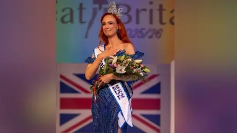 Chris Perfect Alice Cutler wearing a dark blue sparkly dress, a sash with the words: "Miss Great Britain" and holding a bunch of flowers and a crown in her hair. She has red hair and is stood on stage after winning.