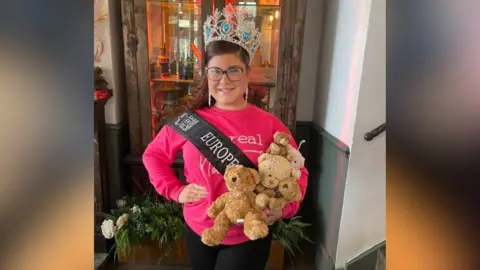 Tracie Steeley Bella is stood wearing her crown and a sash and big silver earrings. She is holding a handful of teddies in one hand and her other hand on her hip. 