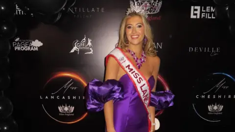 Tabitha Bennett Tabitha stands in front of a black backdrop with small sponsorship logos on it. She wears a purple off the shoulder gown with large, puffy sleeves. She has long blonde hair and wears a silver crown and a red and silver sash. 