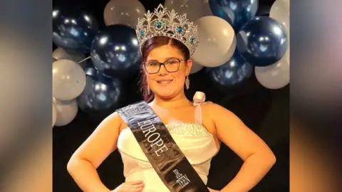 Angela Rowe Chambliss Bella stands on stage wearing crown and a sash and a white dress. She has brown hair tied up and is wearing glasses and silver earrings. A balloon arch is visible in the background.