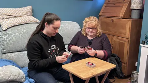 Kate Bradbrook/BBC Victoria and Jane playing a card game. They are sitting on a sofa with a small, wooden card table in front of them. They are both looking at cards they are holding in their hands.