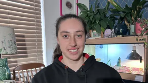 Kate Bradbrook/BBC Victoria, a young woman with dark hair, who is wearing a black hoodie, smiles at the camera. The picture has been taken inside. There is a blind on the left and, behind her, pot plants on top of an aquarium.