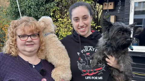 Kate Bradbrook/BBC Jane and Victoria stand next to each other in a garden. Jane, on the left, has a navy blue cardigan and top. She has blonde hair and is wearing glasses. Victoria is wearing a black hoodie. They are both smiling. They are both holding dogs. There is a wooden garden shed with a window behind them.